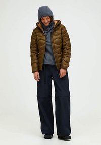 Brun puffig jacka med quiltat mönster, grå tröja och lösa marinblå byxor. Modellen bär en stickad mössa och står framför en neutral bakgrund.