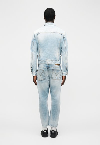 Άνδρας που στέκεται με την πλάτη γυρισμένη, φορώντας ανοιχτό μπλε τζιν μπουφάν και τζιν παντελόνι με μαύρα παπούτσια, σε απλό λευκό φόντο.