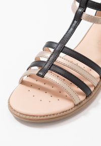 Remsandaler med svarta och beige syntetiska remmar, en vadderad beige sula med perforeringar och en flexibel brun yttersula.