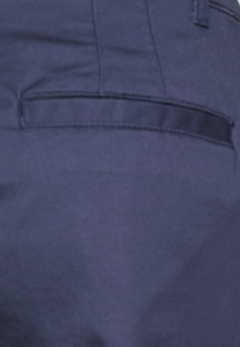 Pantalon bleu marine en tissu texturé, avec une petite poche arrière et un détail de ceinture lisse. Design simple sans motifs visibles.