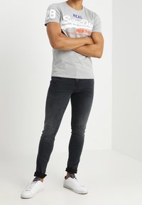 Camiseta gris de manga corta con el logo "SuperDry" y el número 28, combinada con jeans oscuros de corte ajustado y zapatillas blancas, de pie contra un fondo liso.