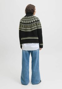 Person mit kurzen braunen Haaren, der einen schwarzen gemusterten Strickpullover, ein weißes Hemd und weit geschnittene blaue Jeans trägt, seitlich vor einem schlichten Hintergrund steht.