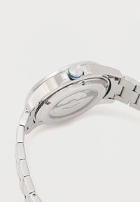 Rozsdamentes acél óra kerek tokban, ezüst szíjjal, kék részletekkel ellátott koronával és világos arcra szerelt egyszerű számlap dizájnnal.