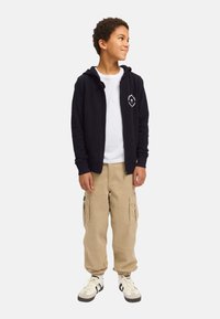 Marineblaue Kapuzenjacke mit Reißverschluss und einer vorderen Tasche, weißes Logo auf der Brust, darüber getragen ein weißes T-Shirt, kombiniert mit beigen Cargo-Hosen und Sneakers.