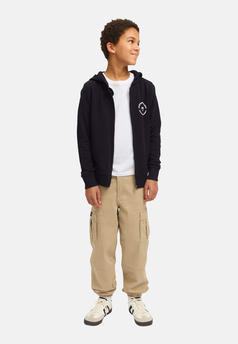 Marineblaue Kapuzenjacke mit Reißverschluss und einer vorderen Tasche, weißes Logo auf der Brust, darüber getragen ein weißes T-Shirt, kombiniert mit beigen Cargo-Hosen und Sneakers.