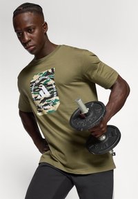 Mand iført en olivengrøn camouflage-logo t-shirt løfter en sort håndvægt med et fokuseret udtryk mod en neutral baggrund.