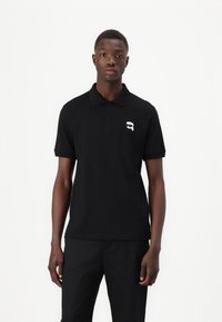 Μαύρο πόλο μπλουζάκι με κοντά μανίκια, τριπλή κουμπότρυπα και μικρό λευκό γραφικό λογότυπο στο αριστερό στήθος. Φτιαγμένο από βαμβάκι.