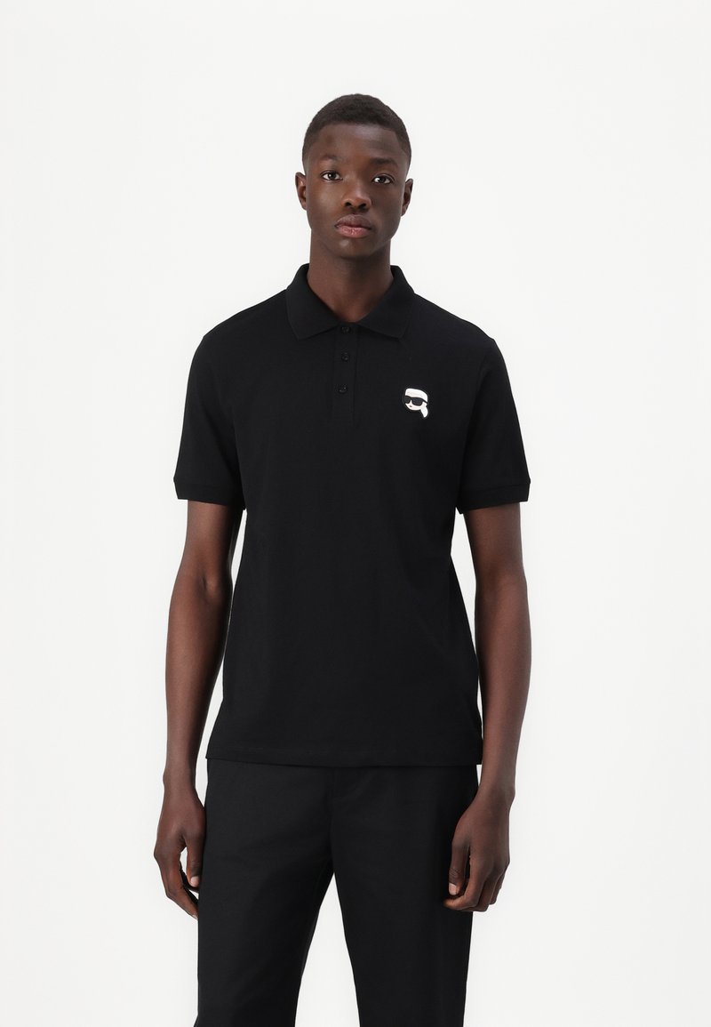 Μαύρο πόλο μπλουζάκι με κοντά μανίκια, τριπλή κουμπότρυπα και μικρό λευκό γραφικό λογότυπο στο αριστερό στήθος. Φτιαγμένο από βαμβάκι.