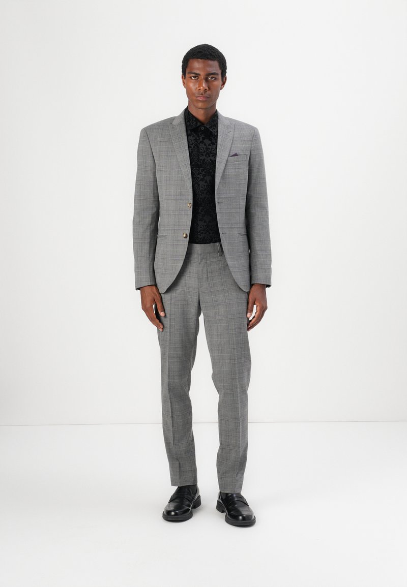 Grijze geruite blazer met notch revers, twee knopen en bijpassende broek, gedragen over een zwart gestructureerd shirt, gecombineerd met zwarte schoenen.