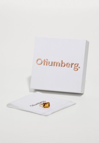 Bague Otiumberg présentée sur un tissu blanc et dans une boîte. La bague comporte une pierre d'ambre rectangulaire et des accents en argent. La boîte possède un logo en relief.