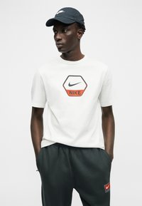 Νέος άνδρας που φοράει λευκό t-shirt Nike με λογότυπο, μαύρο καπέλο Nike και μαύρη φόρμα Nike, στέκεται με τα χέρια στις τσέπες.