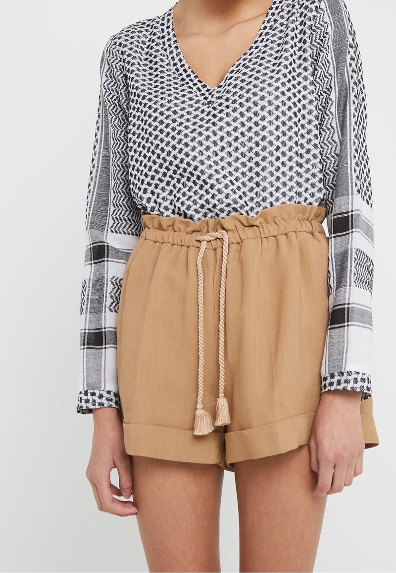Femme portant un short beige à taille haute style paperbag avec une ceinture en corde nouée et un haut à manches longues à col en V à motif noir et blanc.