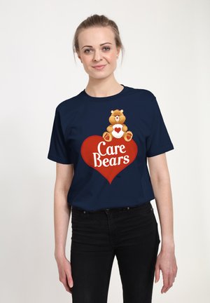 Žena v tmavě modrém tričku s karikaturou medvěda sedícího na červeném srdci a textem "Care Bears" na přední straně, stojící před jednoduchým pozadím.