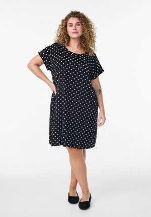 Femme aux cheveux bouclés portant une robe noire à pois blancs longueur genou et des ballerines noires, debout avec une main sur la hanche.