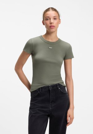Jonge vrouw met lichte huid en haar naar achter gebonden, die een aansluitend olijfgroen kort mouwen shirt en zwarte hoge taille jeans draagt, staand tegen een witte achtergrond.