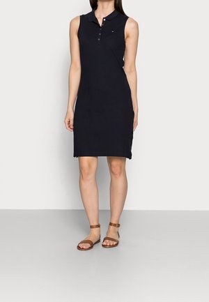 Marineblaues, ärmelloses Polokleid mit Kragen, ausgestattet mit einer Knopfleiste und einem kleinen Logo, kombiniert mit braunen Sandalen mit Knöchelriemen.