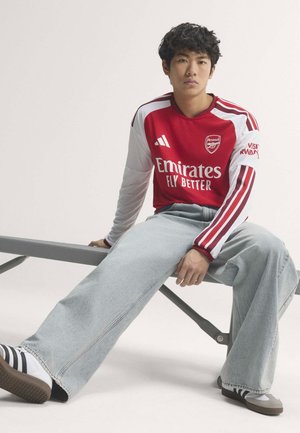 Jeune personne portant un maillot de football Arsenal rouge et blanc, un jean bleu clair à jambes larges, et des baskets rayées, assise sur un banc.