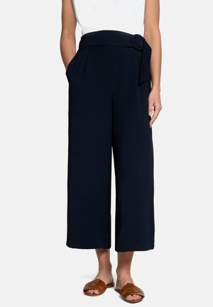 Femme portant un pantalon noir large et court avec une ceinture en tissu et une boucle ronde, associé à des sandales plates marron à bouts ouverts.
