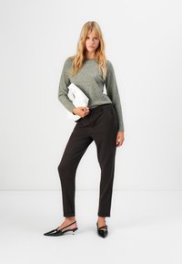 Woman stands against white background wearing gray sweater, dark tapered pants, black slingback heels, holding white folded ice skate.