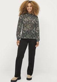 Langmouw blouse in doorschijnende zwarte stof met een bloemenpatroon, gecombineerd met zwarte broeken en beige instappers. Gedetailleerde elastische boorden.