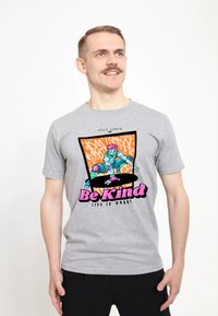 Šedé tričko s grafickým potiskem zobrazující modrou postavu, růžové akcenty a text "Buď hodný" na živě oranžovém pozadí.