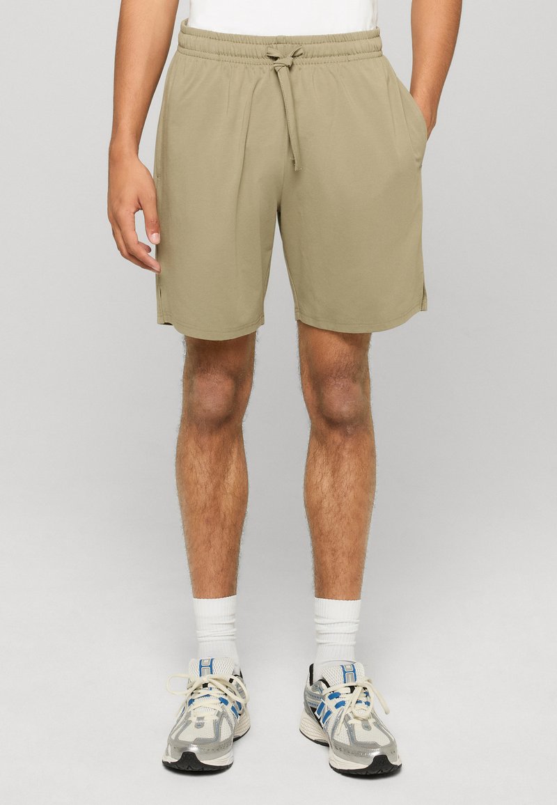 Homme portant un short beige à cordon, des chaussettes blanches montantes et des chaussures de course grises, debout devant un fond uni.