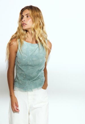 Femme aux longs cheveux blonds portant un haut en dentelle bleu clair sans manches et un pantalon blanc, regardant vers la gauche avec une main dans la poche, sur fond blanc.