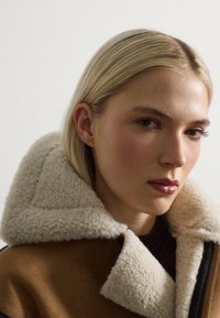 Chaqueta de piel sintética de color marrón con cuello crema, que presenta una superficie texturizada y un cierre con cremallera. El modelo muestra la cara y la parte superior del cuerpo.