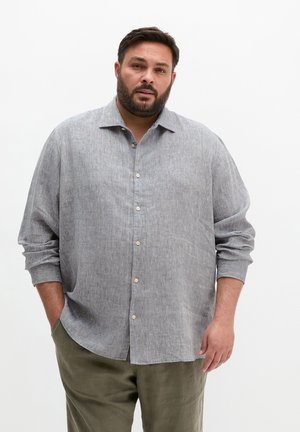 Man with beard wearing a light gray buttoned long-sleeve shirt and olive green pants, standing with one hand in pocket.