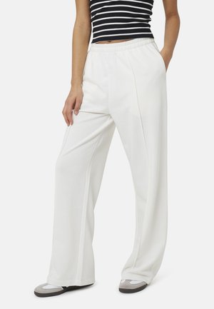 Persoon draagt witte wijde broek, een zwart-wit gestreept mouwloos topje en grijs-witte schoenen, handen in de zakken.