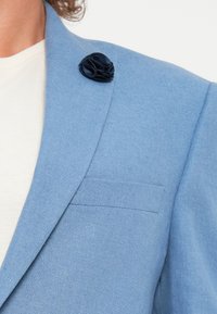 Blazer en lin bleu clair avec une texture subtile, orné d'une broche en forme de fleur navy sur le revers et d'une poche poitrine unique. T-shirt blanc simple en dessous.