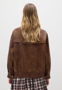Persona con lunghi capelli biondi ondulati che indossa una giacca di pelle marrone e una gonna a quadri, in piedi su uno sfondo bianco semplice.