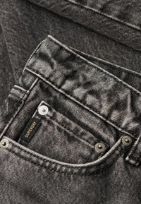 Los vaqueros negros de denim cuentan con un bolsillo frontal de forma rectangular, botones de hardware plateados y una etiqueta con el nombre de la marca "Superdry".