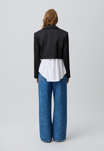 Blazer negro recortado con solapas anchas, usado sobre una camisa blanca, combinado con unos pantalones vaqueros de denim azul claro de corte holgado.