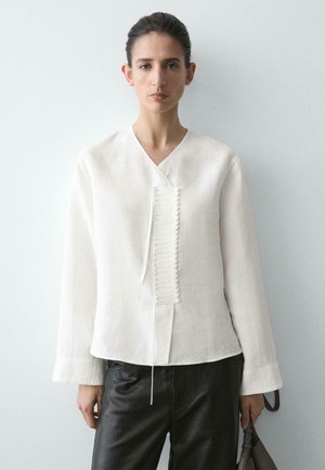 Femme aux cheveux foncés portant une chemise blanche à manches longues avec détail texturé de boutons, pantalon noir, tenant un sac sombre devant un fond clair uni.