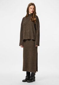 Femme portant un pull en tricot côtelé marron et une jupe longue assortie avec une fente sur le côté, accompagnée de bottines noires à boucles, se tenant devant un fond blanc.