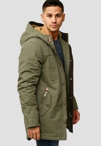 Oliven grønn parka med pelsforet hette, med dobbelt glidelås og knappelukking, sidelommer med glidelås og kontrasterende brune detaljer.