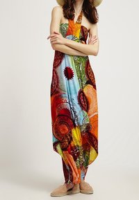 Femme portant une robe maxi à col halter colorée et à motifs, debout avec les bras croisés, associée à des chaussures slip-on beiges.