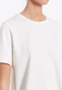 T-shirt bianca realizzata in tessuto morbido, con scollo rotondo, maniche corte, dalla texture liscia e design minimalista, che mostra un primo piano della spalla.