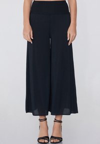 Pantalons larges noirs avec taille smockée, fabriqués en tissu léger. Associés à des sandales noires à lanières, dévoilant les chevilles.