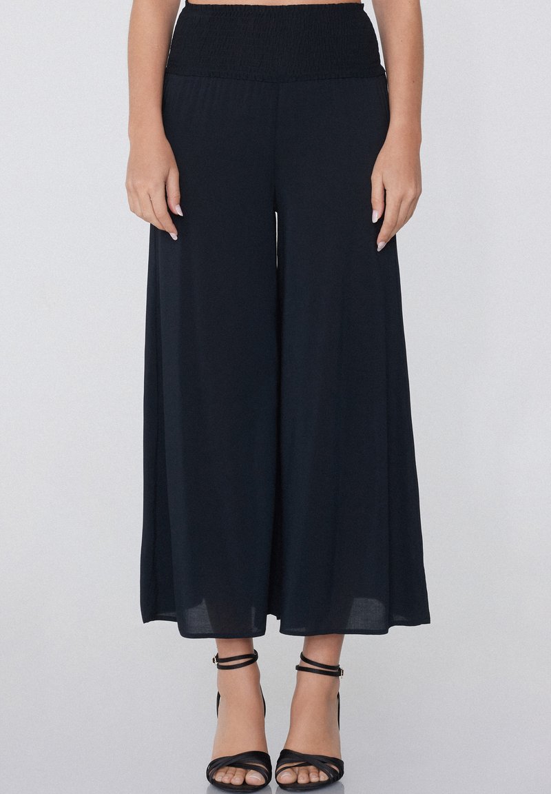 Pantalons larges noirs avec taille smockée, fabriqués en tissu léger. Associés à des sandales noires à lanières, dévoilant les chevilles.