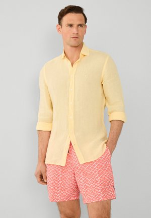 Camisa de lino amarilla clara con mangas arremangadas, combinada con pantalones cortos rosas que presentan un patrón de conchas blancas. Logo sutil en la camisa.