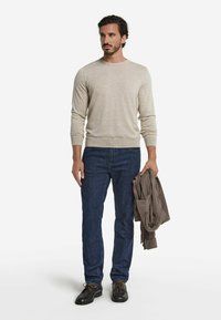 Beige långärmad tröja, mörkblå jeans och svarta skor. Personen håller en brun tröja över armen. Bomullstyg, avslappnad design.