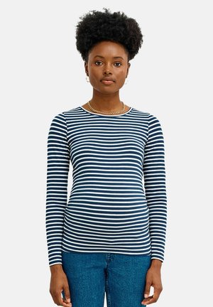 Frau mit natürlichem lockigem Haar, die ein marineblau-weiß gestreiftes Langarmshirt und blaue Jeans trägt und vor einem einfarbigen Hintergrund steht.