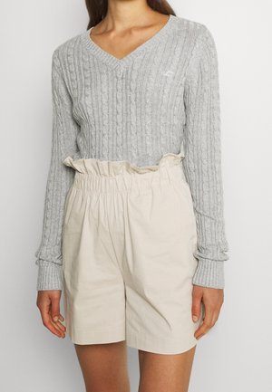 Vrouw draagt een lichtgrijze kabelgebreide V-hals trui en beige hoog getailleerde paperbag shorts, staand tegen een effen achtergrond.
