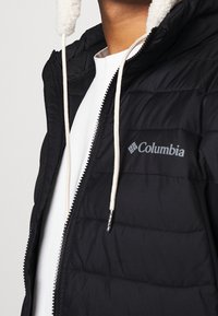 Svart puffjacka med dragkedja, vit fleece-fodrad huva och Columbia-logotyp på bröstet. Har en quiltad textur och kontrasterande dragsnören.