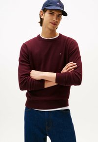 Suéter burdeos con cuello redondo y puños acanalados, usado sobre una camisa blanca. Vaqueros de mezclilla con costuras visibles. Modelo de pie con los brazos cruzados.