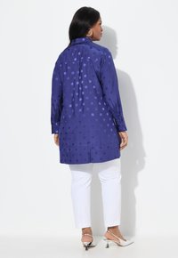 Blusa de manga larga abotonada en azul profundo con lunares en relieve. Presenta un respaldo plisado con un dobladillo ligeramente más largo. Lleva puesta con pantalones blancos.