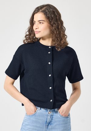 Mujer con cabello rizado y castaño que lleva una blusa negra de manga corta con botones y vaqueros azul claro, con las manos en los bolsillos, mirando hacia un lado.