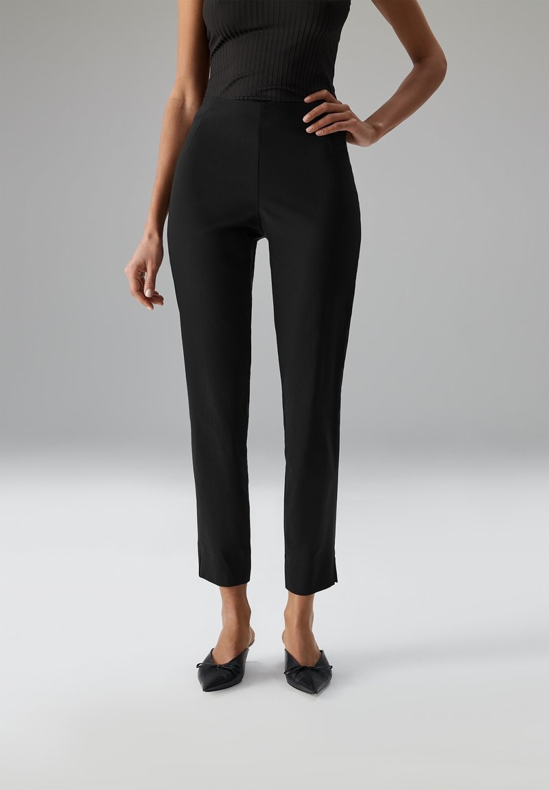 Vrouw in een strakke zwarte enkel-lange broek, zwarte mouwloze top en zwarte puntige hakken, staand met één hand op de heup.